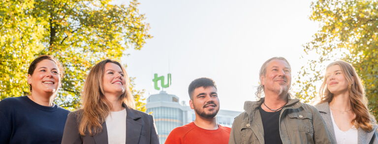 Eine Gruppe von Personen läuft über den Campus der TU Dortmund. Im Hintergrund ist der Mathetower mit dem TU Logo zu sehen. Die Bäume sind grün und die Sonne scheint.