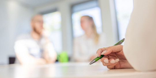 Nahaufnahme einer Stift haltenden Frauenhand, die auf einem Tisch abgelegt ist. Verschwommen im Hintergrund sind zwei weitere Personen im Gespräch zu sehen.