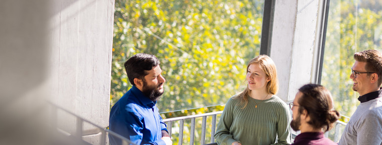 Gespräch im Treppenhaus Vier Personen stehen vor einem Fenster im Treppenhaus eines Universitätsgebäudes und unterhalten sich.