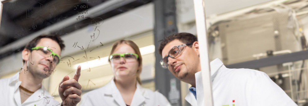 Drei Personen mit weißem Kittel und Schutzbrille stehen im Labor vor einem transparenten Board und betrachten eine chemische Formel.