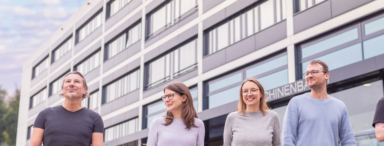 Fünf Beschäftigte der TU Dortmund laufen über den Campus. Im Hintergrund steht das Gebäude Maschinenbau.