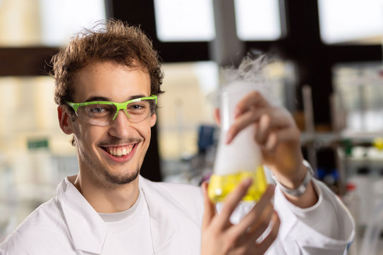 Auszubildender Luca trägt Schutzbrille und Laborkittel und hat einen Glaskolben mit einer gelben, dampfenden Flüssigkeit in der Hand.
