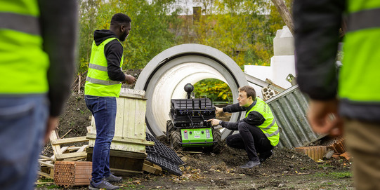 Mitarbeiter in gelben Westen hantieren mit einem Roboter auf Rädern, der draußen auf einem Testgelände vor einer Betonröhre steht.