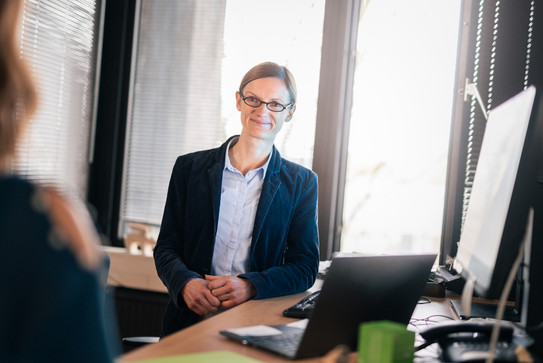 Dr. Kathrin Höhner lehnt an ihrem Schreibtisch und wendet sich einer weiteren Person zu.