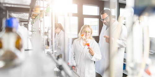 Personen mit weißem Kittel und Schutzbrille im Labor. Eine junge Frau zeigt einem Mann eine Glasgefäß mit einer orangefarbenen Flüssigkeit.