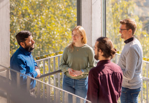 Vier Personen stehen vor einem Fenster im Treppenhaus eines Universitätsgebäudes und unterhalten sich.
